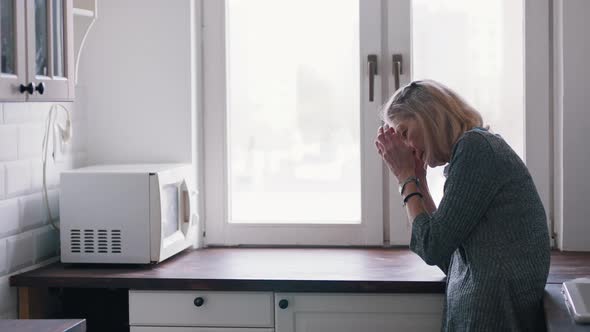 Sorrowful Lonely Old Woman Leaning on the Kitchen Counter and Looking Through the Window alt