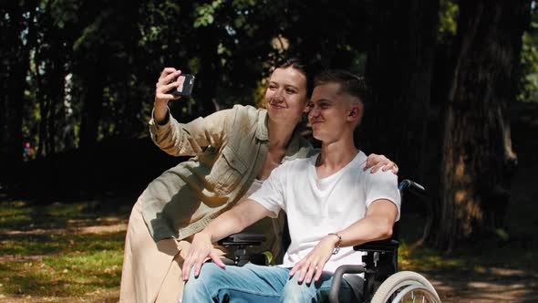 Man in a Wheelchair and His Girlfriend Spending Time at the Summer Park and Taking a Selfie Together alt