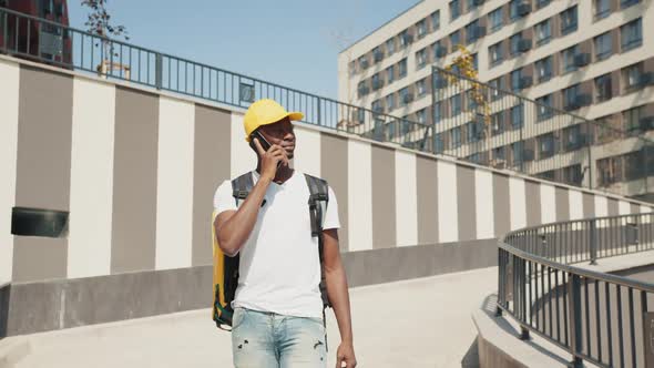 A Young AfricanAmerican Delivery Man Walks Down a City Street with a Yellow alt