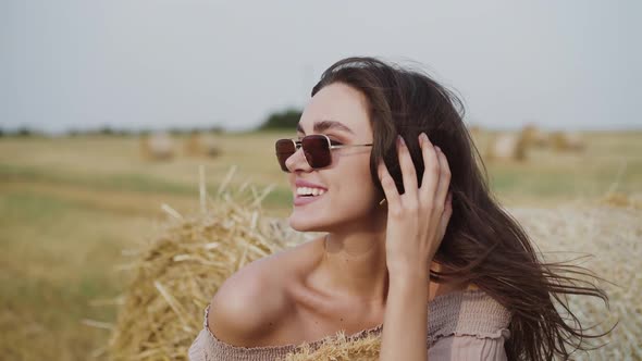 Happy Woman Rejoices at Haystack and Corrects Hair During Strong Wind in Field alt