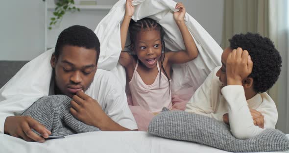 Busy Father Lies with His Wife Under Covers Looking at Mobile Phone Suddenly Funny Daughter Appears alt