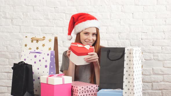 Cheerful Red-haired Girl in Santa's Hat Holds a Christmas Box and Smiles at the Camera.