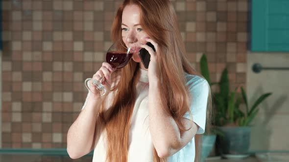 young red-haired woman with glass of red wine in kitchen talking on phone alt