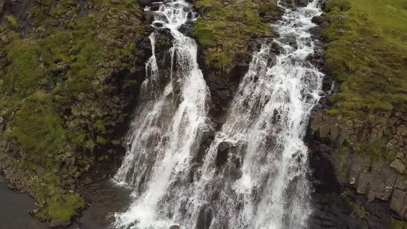 Aerial rising over Glymur waterfalls and river streaming down rocky cliff surrounded by verdant high alt