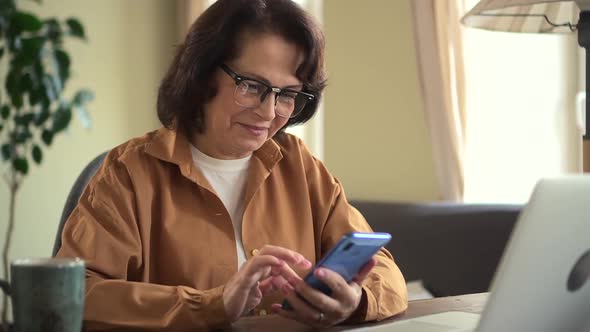 Ederly Woman Using Phone and Sitting at Table with Laptop in Apartment Room Indoors alt