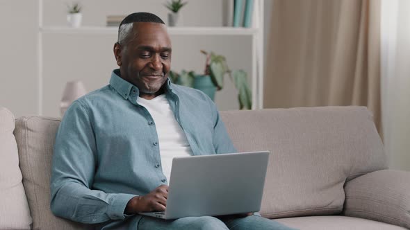 Mature Happy African American Man Sitting on Sofa at Home Relaxing Smiling Chatting on Laptop Via alt