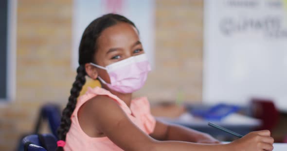 Portrait of mixed race schoolgirl wearing face mask, sitting in classroom looking at camera alt