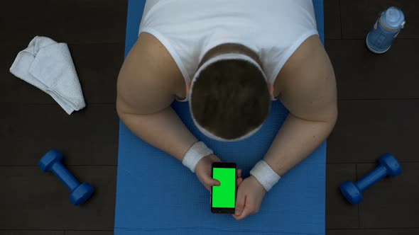 Plump Male Using Online Fitness Training Application, Lying With Smartphone Mat alt