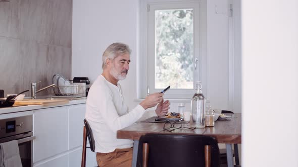 Mature man using smartphone at breakfast table alt