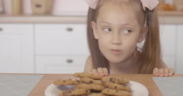 Cheerful Little Girl Peeking Out of Table and Taking Sweet Tasty Cookie From Plate alt