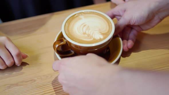 Barista's Hands Put a Brown Cappuccino Mug with Beautiful Latte Art on the Table Where Female Hands alt