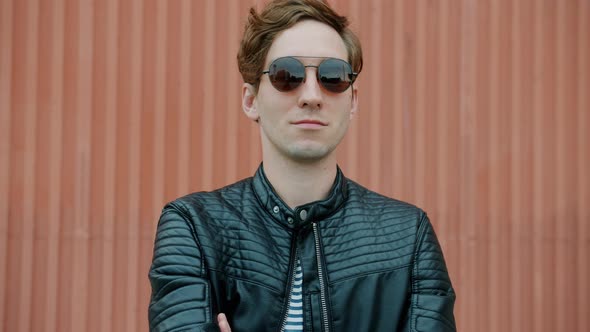 Portrait of Cool Guy Wearing Sunglasses and Leather Jacket Standing Outdoors with Arms Crossed alt