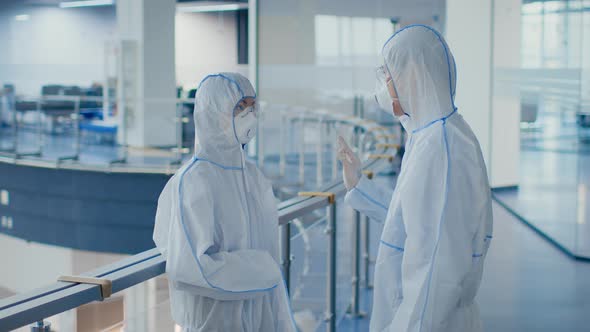 Two Laboratory Workers Talking Wearing Hazmat Suits And Masks Indoors alt
