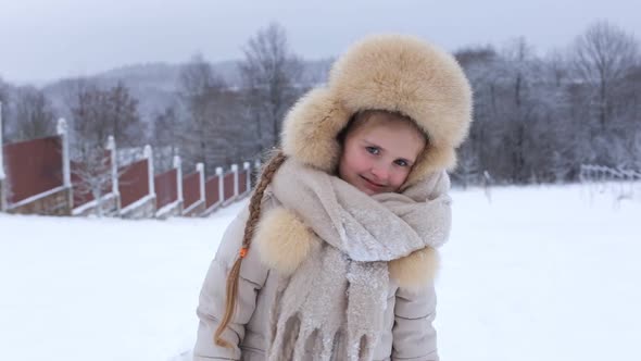 A Beautiful Little European Girl in a Fur Hat Looks at the Camera and Smiles