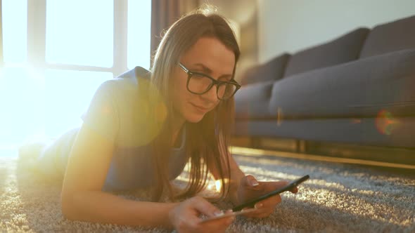 Happy Woman with Glasses Is Lying on the Floor and Makes an Online Purchase Using a Credit Card and alt
