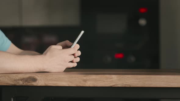A Man with a Mobile Phone in His Hands Stands Near a Wooden Table Uses a Mobile Application alt