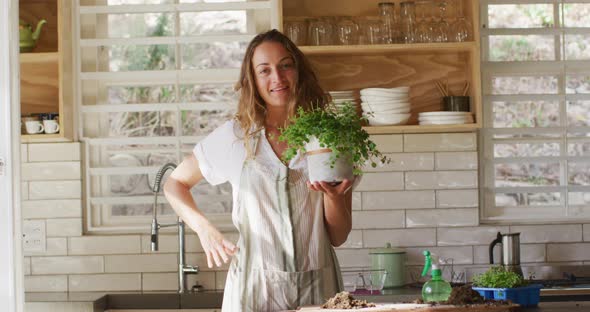 Portrait of smiling caucasian woman holding potted plant, standing in sunny cottage kitchen alt
