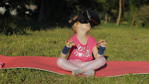 Child in VR Headset Helmet Sitting in Lotus Position on Mat and Performing Yoga Meditation in Park alt