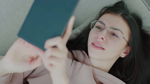 Young woman with glasses using mobile device. Girl tapping and swiping on tablet lying on sofa