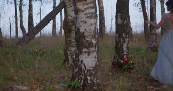 A Bride in a Gray Dress Walks with a Wedding Bouquet in the Forest alt