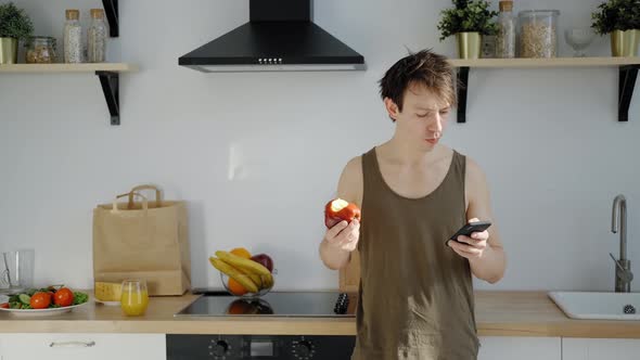 Man in a Sleeveless T-shirt Eating a Red Apple, Looking at Smartphone