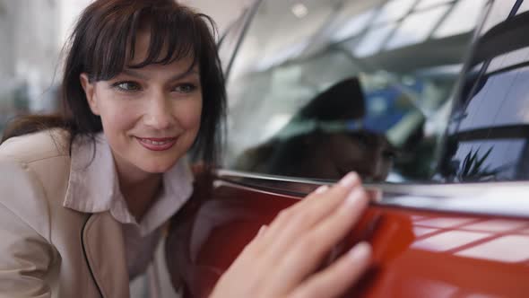 Excited Caucasian Woman Touching Brand New Red Car in Dealership Smiling alt