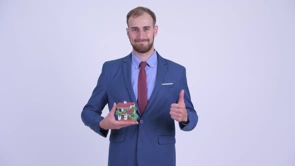 Happy Bearded Businessman Holding House Figurine and Giving Thumbs Up