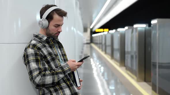 Young Man Waits for a Train in the Subway Uses a Smartphone and Listens to Music with Wireless alt
