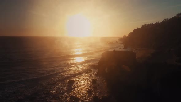 Drone rising over rocks of El Matador Beach to the bright setting sun in Malibu, Califronia, USA alt