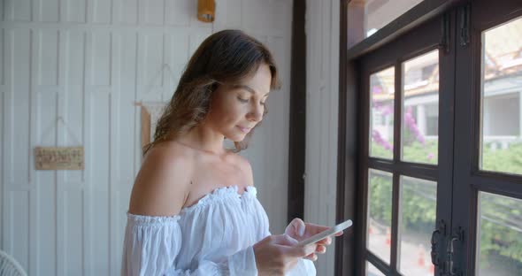 Brunette Woman Stands By the Window Indoors and Types the Text Massage and Looking to the Window alt