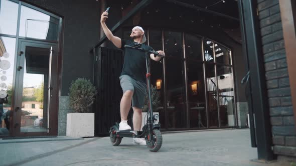 Young Man Standing with Escooter in City Downtown and Taking Selfie with Smartphone alt