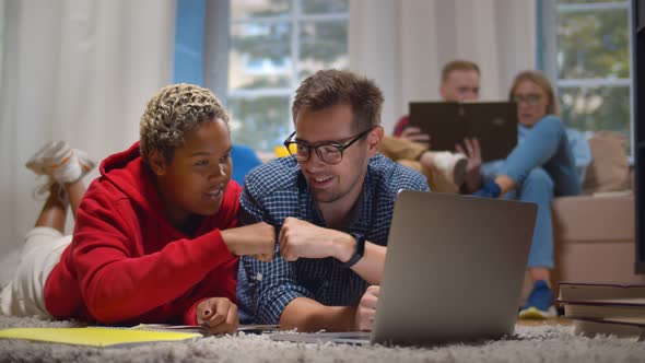 Diverse Student Working on Project Together Gesturing Fist Bump Lying on Floor in Dorm Using Laptop alt