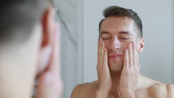 Close-up Portrait of Young Man Applying Cream on Face Looking in Mirror. Handsome Young Male alt