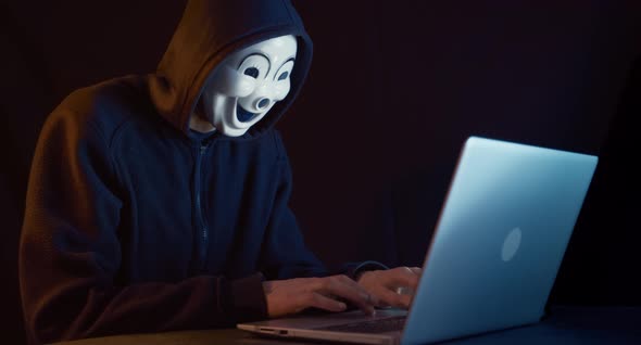Hacker with masked face uses laptop to hack data. alt