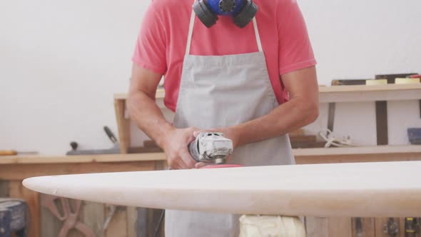 Caucasian male surfboard maker wearing a face mask and preparing to polishing a surfboard alt