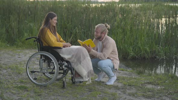 Wide Shot of Loving Caucasian Man Reading for Paralyzed Woman at Sunset on River Bank. Portrait of alt
