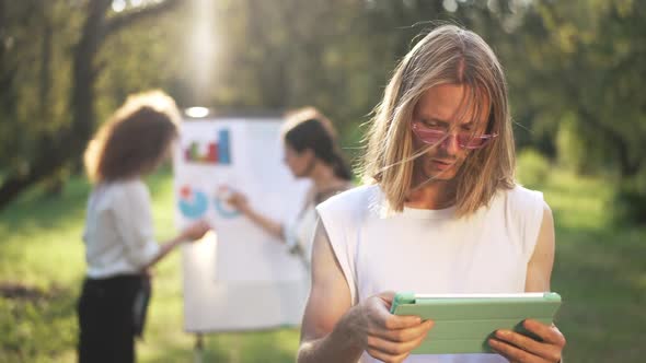 Portrait of Thoughtful Concentrated Young Man Surfing Internet on Tablet with Blurred Women alt