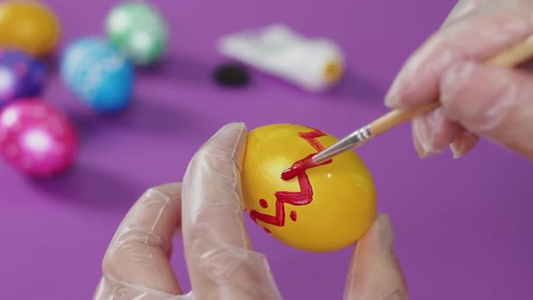 A man's hand, wearing a transparent glove, draws red dots on a Easter egg on a purple table.