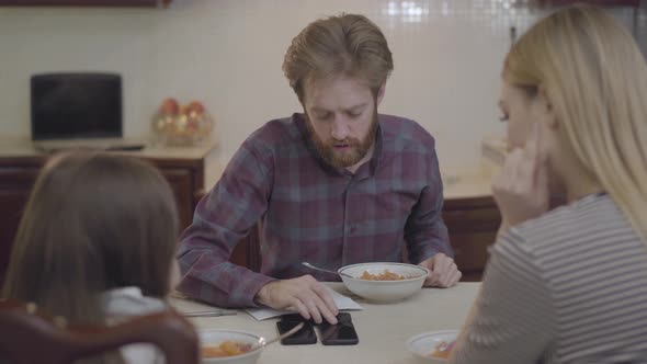 The Family of Three People Having Breakfast Sitting at the Table. Busy Father Talking By Cell Phone alt