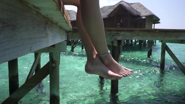 Young lady sitting on wooden jetty. Crossed legs swinging above turquoise ocean. Maldives. Bungalow alt
