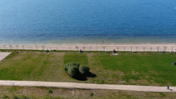 Lake beach with people walking and sunbathing and bathing, lawn garden with shrubs. Overhead drone s alt