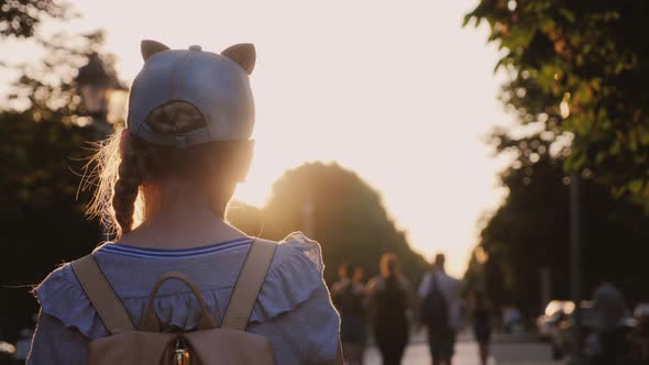 A Girl with a Backpack Is in a City Park, Passersby Go By alt