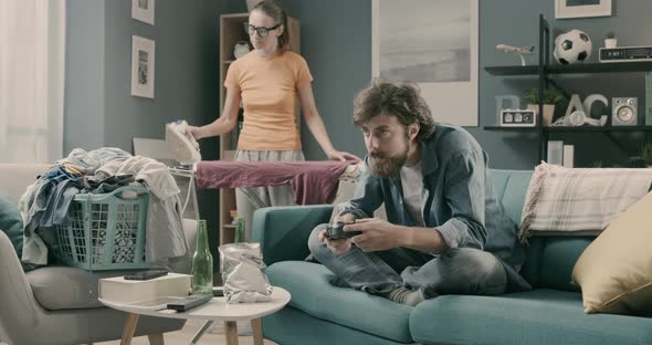 Lazy husband playing videogames while his wife is ironing