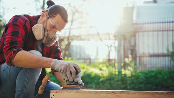 A Young Man in a Construction Respirator Grinds a Parquet Board with a Sander alt