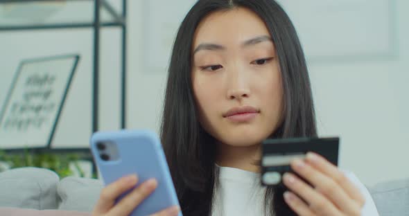 Young Smiling Asian Woman Shopping Online Holding Smart Phone and Credit Card alt