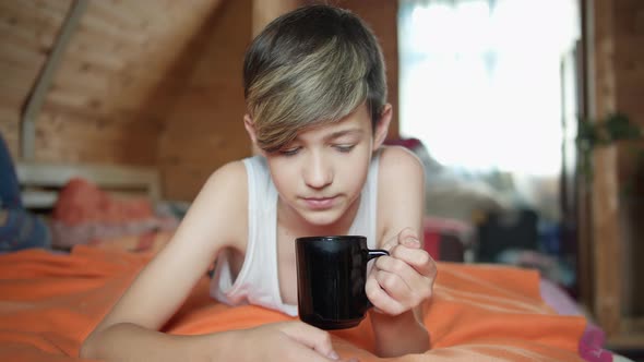 Boy Lies on the Bed Drinking Hot Tea and Smiling Thinking About Something