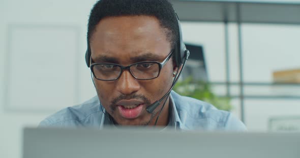 Portrait of African Man in Glasses Working Online Video Conference Using a Laptop with Headset at alt
