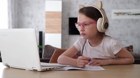 A Smiling Little Caucasian Girl with Headphones Looks at Her Laptop Sitting at the Table Listening