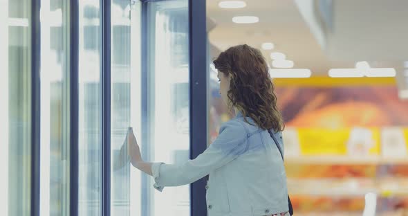 Pretty Girl Opens a Refrigerated Display Case in a Store and Takes Out a Bag of Frozen Vegetables alt