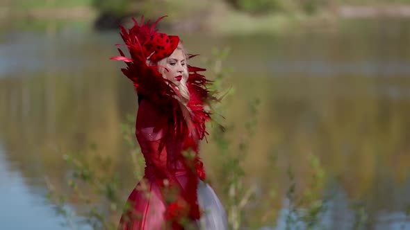 Portrait of a Blonde in a Red Dress with a Feather Collar and a Hat on the Background of a Blurred alt